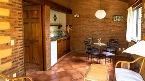 A rustic kitchen with a brick wall, wooden cabinets, and a dining area with a table and chairs.