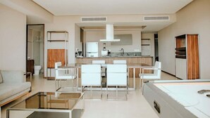 Ett modernt hotellrum med ett litet kök, en matplats med vita stolar, en soffa och ett soffbord i glas.