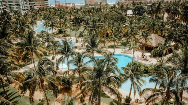 Ett semesterkomplex med flera pooler, omgivet av palmer, en strand och utsikt över närliggande byggnader.