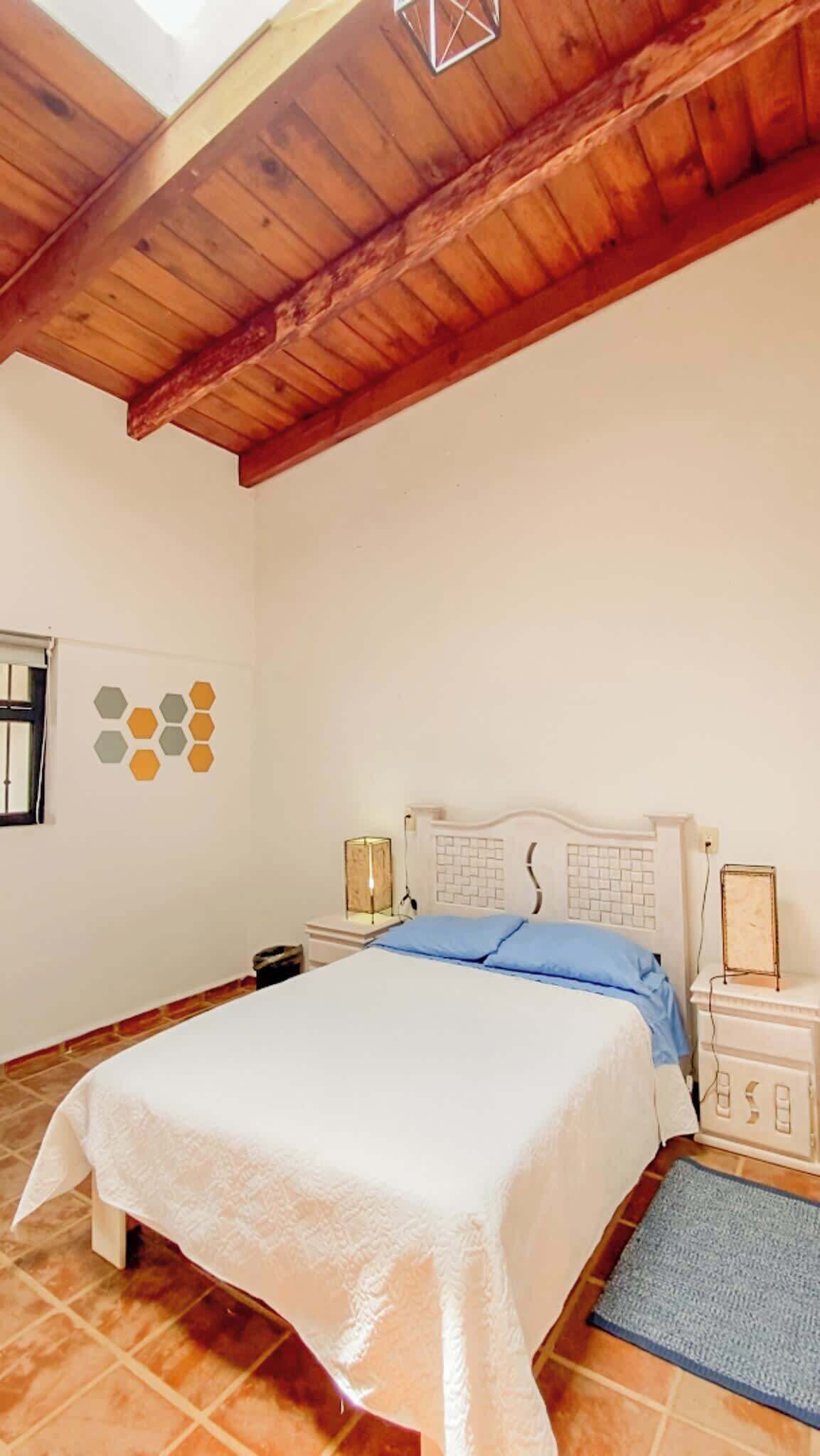A bedroom with a wooden ceiling, a bed with a white bedspread, a nightstand, a bedside table, and a wall decoration.