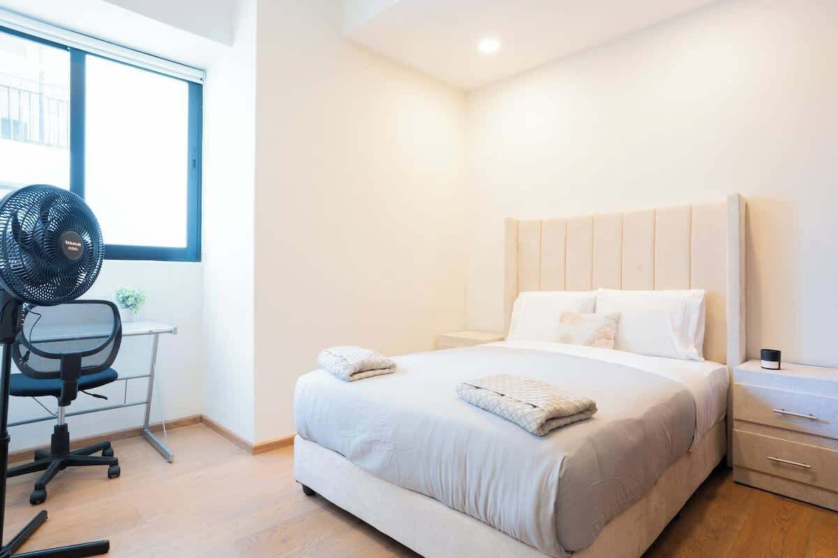 A bedroom with a bed, bedside tables, a desk with a chair, and a standing fan.