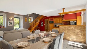 Un coin repas avec une table en bois dressée pour quatre personnes, un escalier et une cuisine avec un mur de crédence en briques.