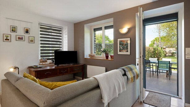 Un salon comprenant un canapé, un téléviseur posé sur un meuble en bois, une fenêtre avec des stores et une porte donnant sur un espace extérieur.