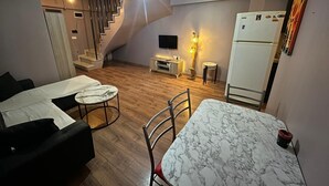 Ein kompakter Wohnraum mit einer Couch, einem Esstisch, einem Fernseher, einem Kühlschrank, einer Treppe und einer Lampe.