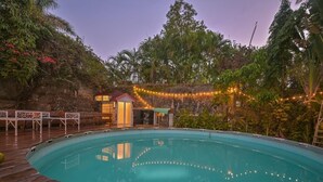 A swimming pool with a wooden deck, surrounded by lush greenery and string lights.