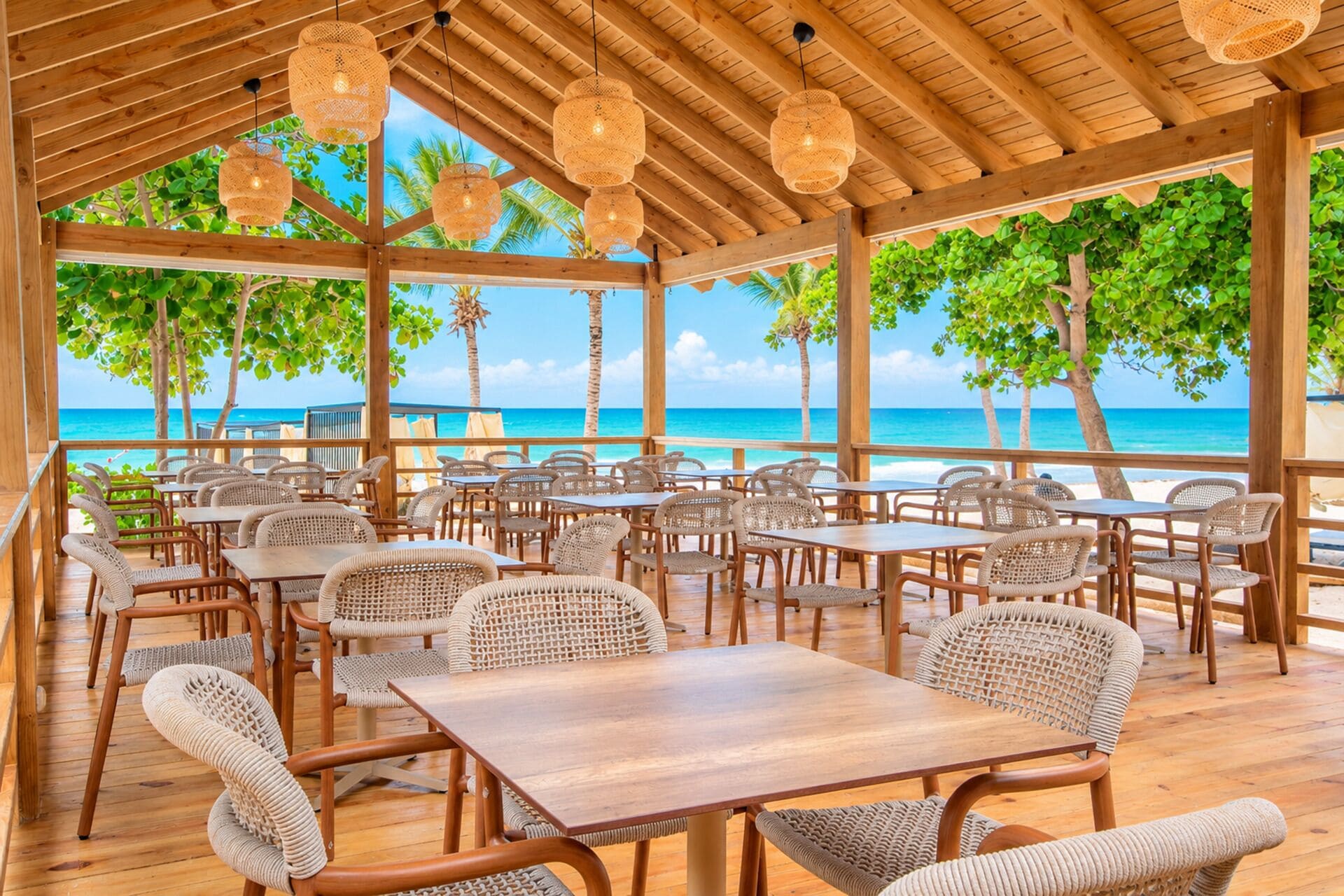 A covered outdoor seating area with tables and chairs, overlooking a beach with palm trees and the ocean.