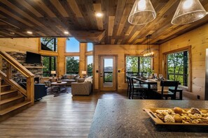 A spacious wooden interior with a stone fireplace, a dining area, and a kitchen counter with bread. - Lexy Lane Indoor Pool | Mountain Views | Sleeps 12 (Sevierville)