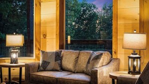 Ein gemütliches Zimmer mit einem Ledersofa, zwei Beistelltischen mit Lampen und Blick auf Bäume durch große Fenster.
