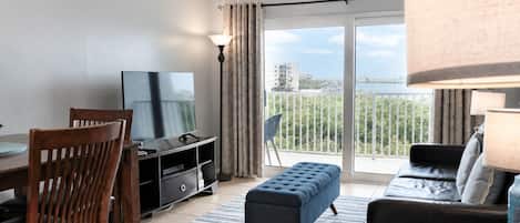 A modern living room with a flat-screen TV, a dining table with chairs, a sofa, and a balcony with a view.
