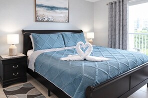 Ein Bett mit blauer Bettdecke, zwei Nachttischlampen, ein schwarzer Nachttisch, ein dekorativer Teppich und ein Gemälde an der Wand.