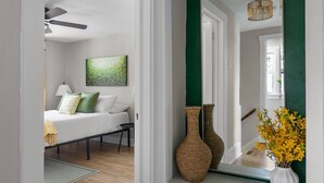 Un pasillo con una cama, dos jarrones de mimbre, un jarrón blanco con flores amarillas y una pared verde decorativa.