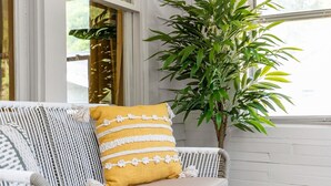 Un sofá de mimbre blanco con un cojín mostaza, una planta en maceta y una alfombra estampada en una habitación con paredes blancas y una ventana.