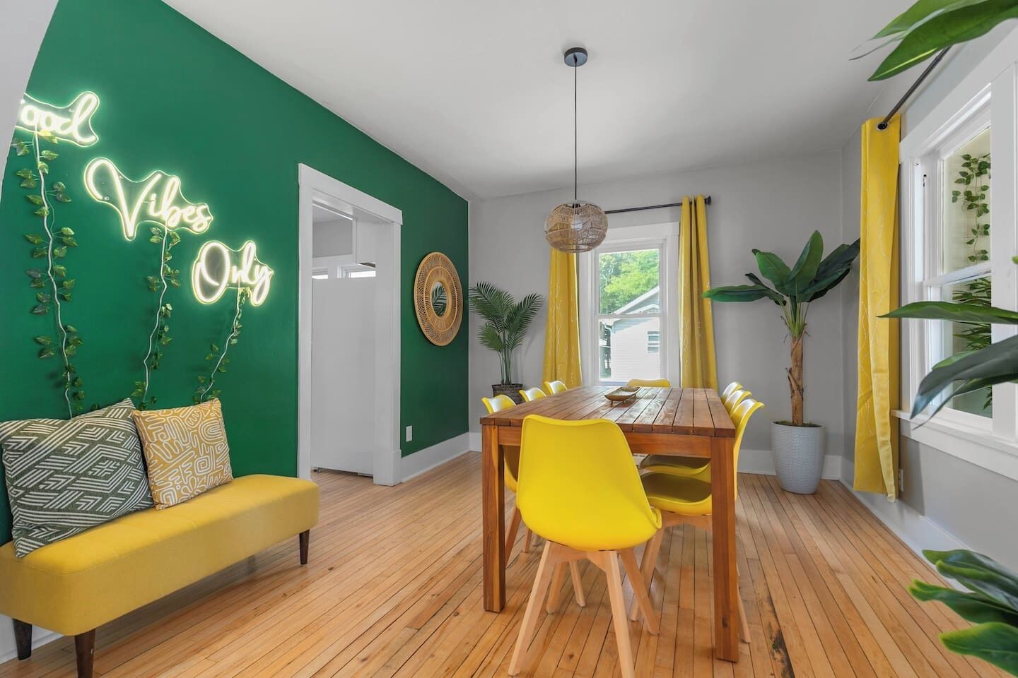 Une pièce avec un mur vert, un panneau lumineux au néon, une table à manger en bois avec des chaises jaunes, un canapé jaune et une plante en pot.