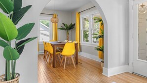 Comedor con mesa de madera y sillas amarillas, rodeado de ventanas amplias y plantas en macetas.