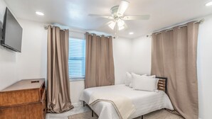 A bedroom with a bed, a wooden dresser, a ceiling fan, and a window with curtains. - Noel Real Estate (Naples)