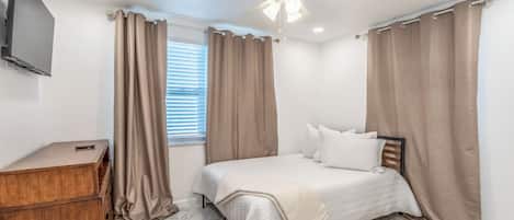 Une chambre à coucher avec un lit, une commode en bois, un ventilateur de plafond et une fenêtre avec des rideaux.