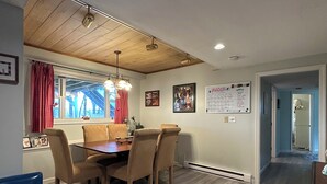 A dining area with a wooden table, four chairs, a blue bench, and a window with red curtains.