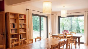 A dining area with a wooden table and chairs, a bookshelf, and large windows with curtains. - La Hulotte en Brocéliande - Yoga, seminars & group holidays 15 pers. (Saint-Malon-sur-Mel)