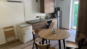 Cocina compacta con refrigerador, microondas y lavaplatos. Hay una mesa redonda de madera con sillas y un ventanal que proporciona luz natural.