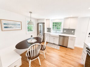 A modern kitchen with white cabinetry, a stainless steel refrigerator, a dining area with a dark table and white chairs, and a window with a view of greenery. - 3-bedroom, 2 bath Popponesset Cottage with AC - walk to beach and The Spit (Mashpee)