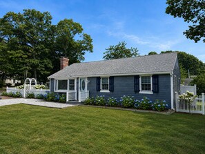 A single-story house with a front porch, a small garden with blue flowers, and a white picket fence. - 3-bedroom, 2 bath Popponesset Cottage with AC - walk to beach and The Spit (Mashpee)