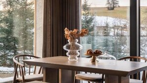 Een eetgedeelte met een houten tafel, twee hanglampen en zicht op een besneeerd landschap door grote ramen.
