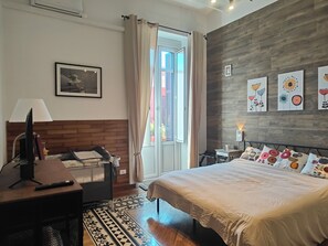 A bedroom with a bed, a desk, a chair, a TV, a window with curtains, and artwork on the wall. - Amapola House Bari (Bari)