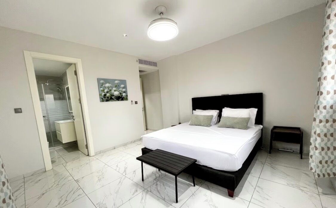 A bedroom with a bed, bedside tables, a bench, a door leading to a bathroom, and a ceiling light.