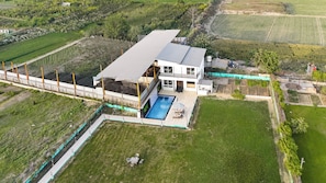 A modern two-story house with a swimming pool, surrounded by a fenced garden and fields. - Unique Farmhouse by ELIVAAS (Noida)