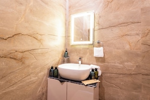 A bathroom with a marble wall, a white sink, a mirror, and toiletries. - Unique Farmhouse by ELIVAAS (Noida)
