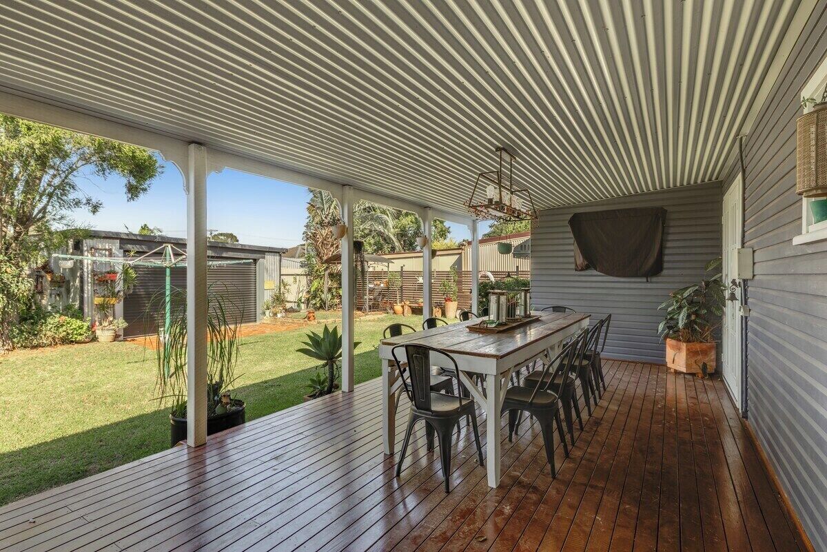 A covered outdoor patio with a wooden table and chairs, surrounded by potted plants and a view of the backyard.