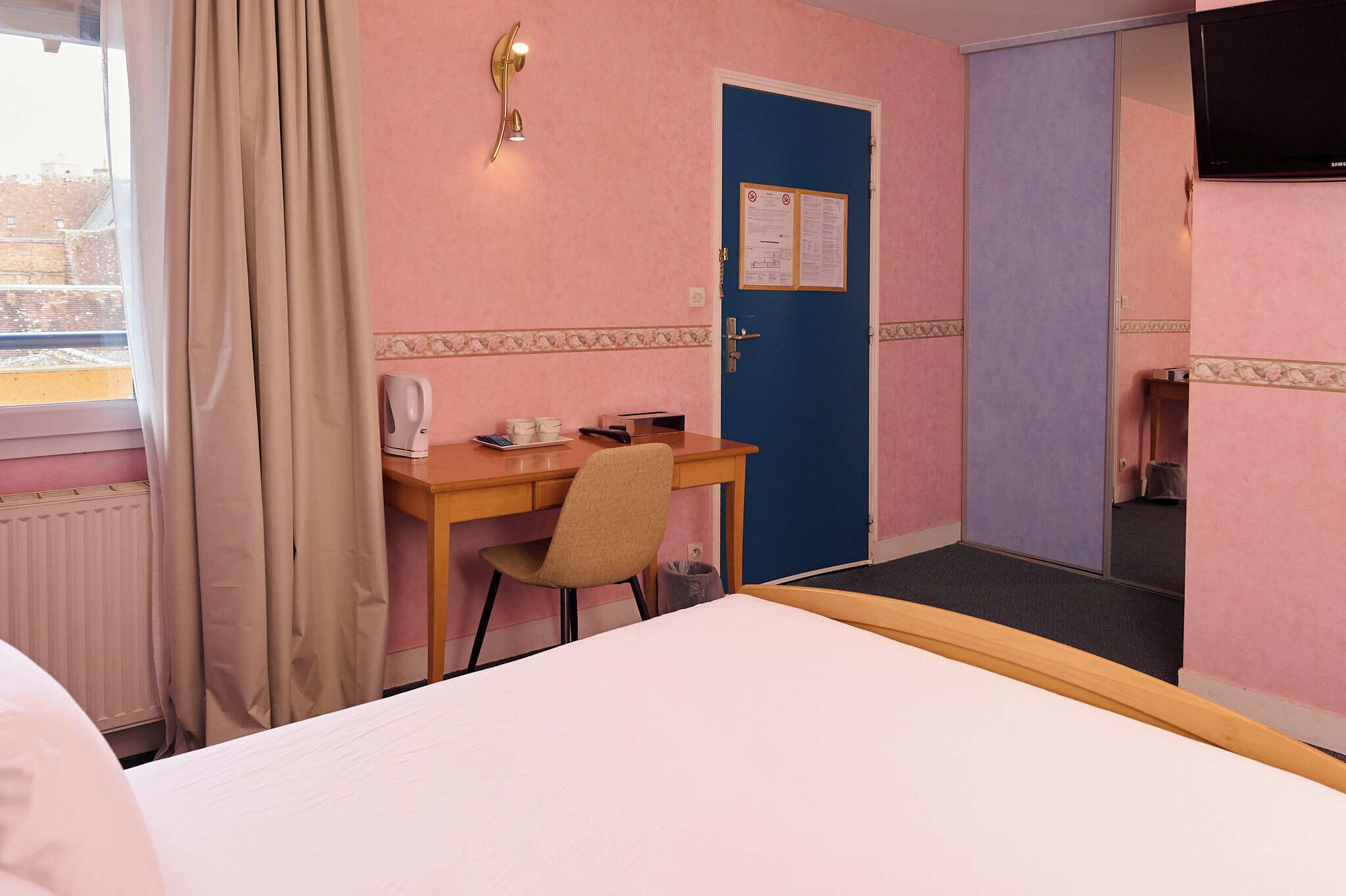 Une chambre d’hôtel avec un lit, un bureau et une chaise, une porte bleue et une fenêtre avec des rideaux.