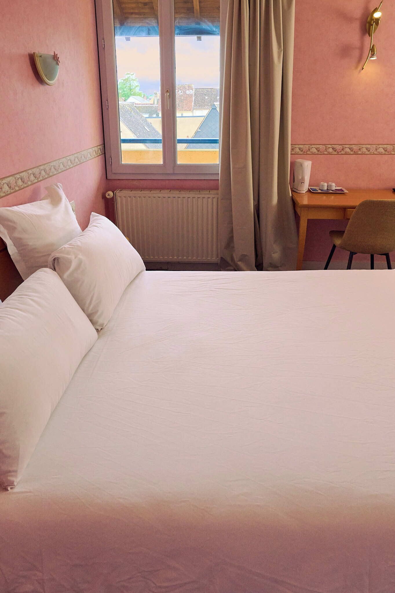 Habitación de hotel con cama, ventana con cortinas, radiador, una mesita con silla y lámpara fijada a la pared.