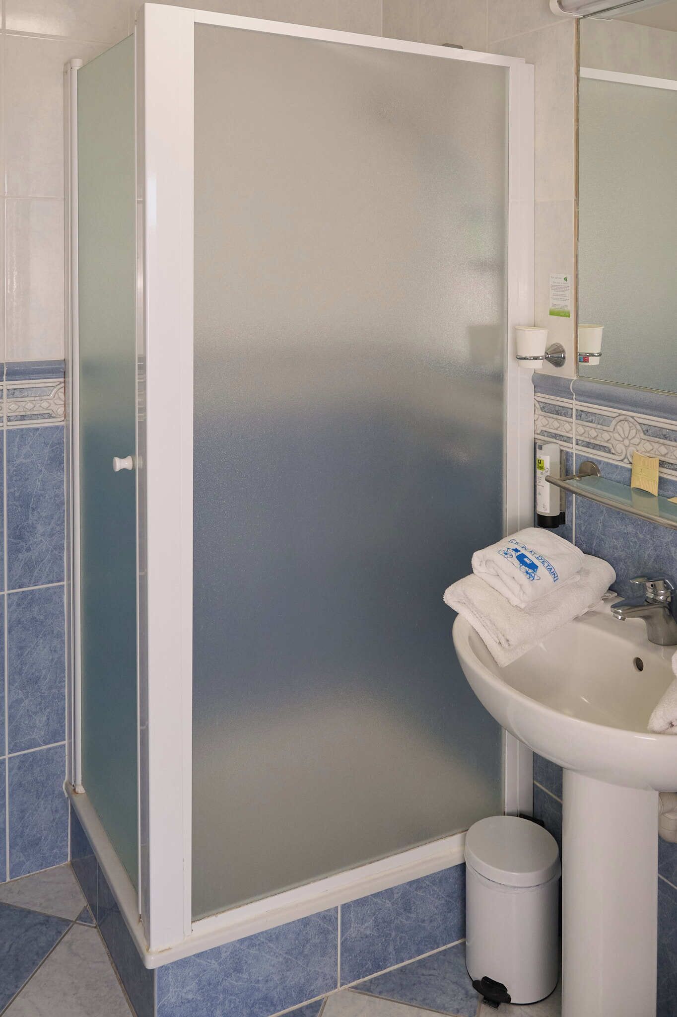 A bathroom with a glass shower enclosure, a sink, a trash bin, and a mirror with a towel on it.