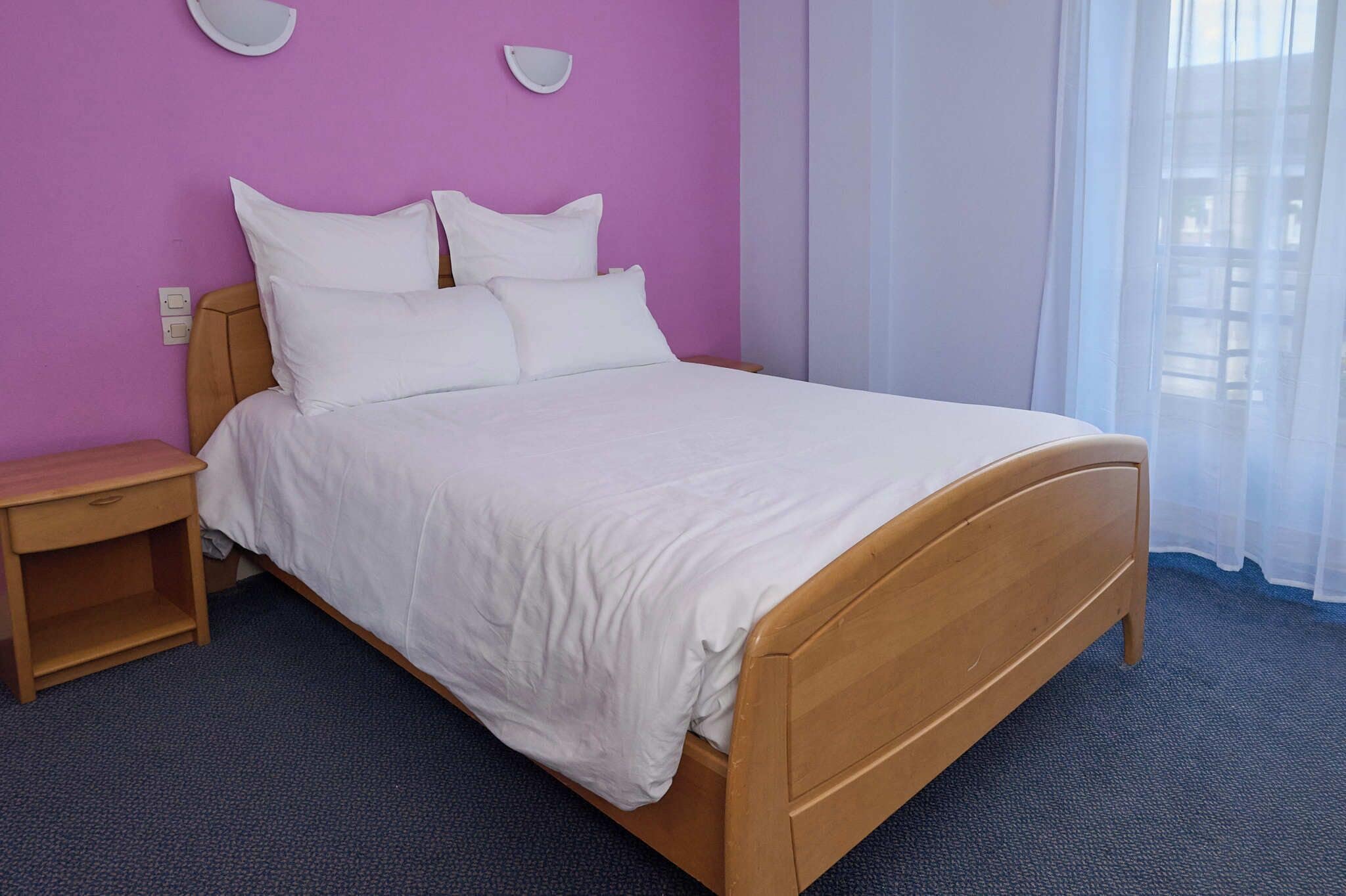 A single bed with a wooden frame and white bedding, a wooden nightstand, and a wall-mounted light fixture.
