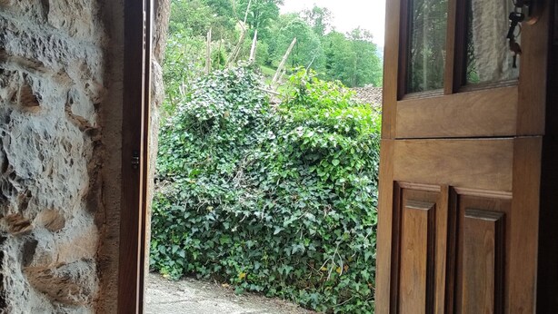 Uma porta de madeira rústica dá para uma área pavimentada, com um muro de pedra de um lado.