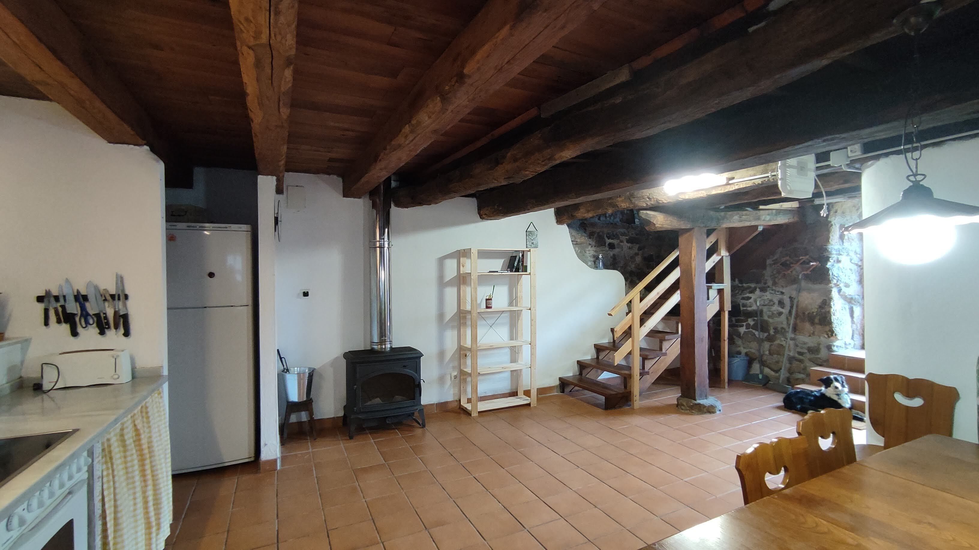 A rustic kitchen with a wooden ceiling, a white refrigerator, a stove, and a wooden staircase leading to a loft.