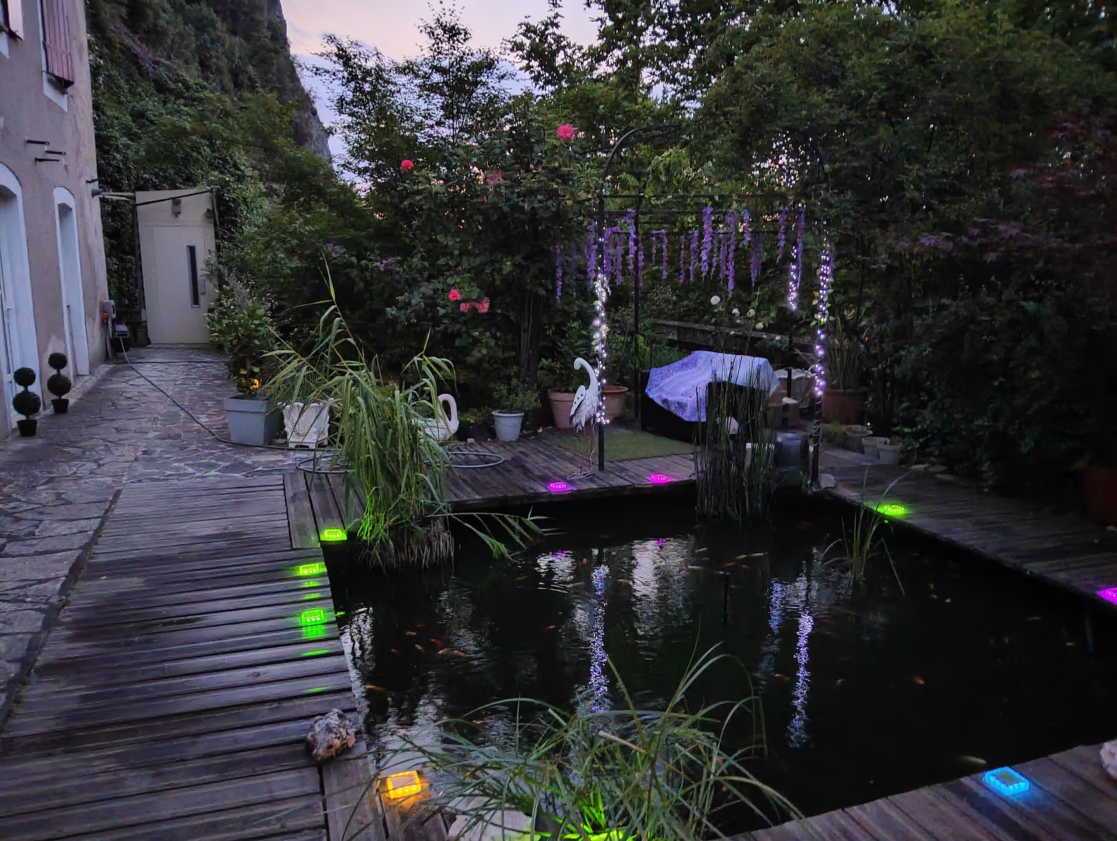 Una terraza de madera que da a un estanque con luces decorativas, rodeada de plantas en macetas y una estatua.