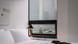 A hotel room with a bed, a nightstand, a lamp, and a window with a view of tall buildings.
