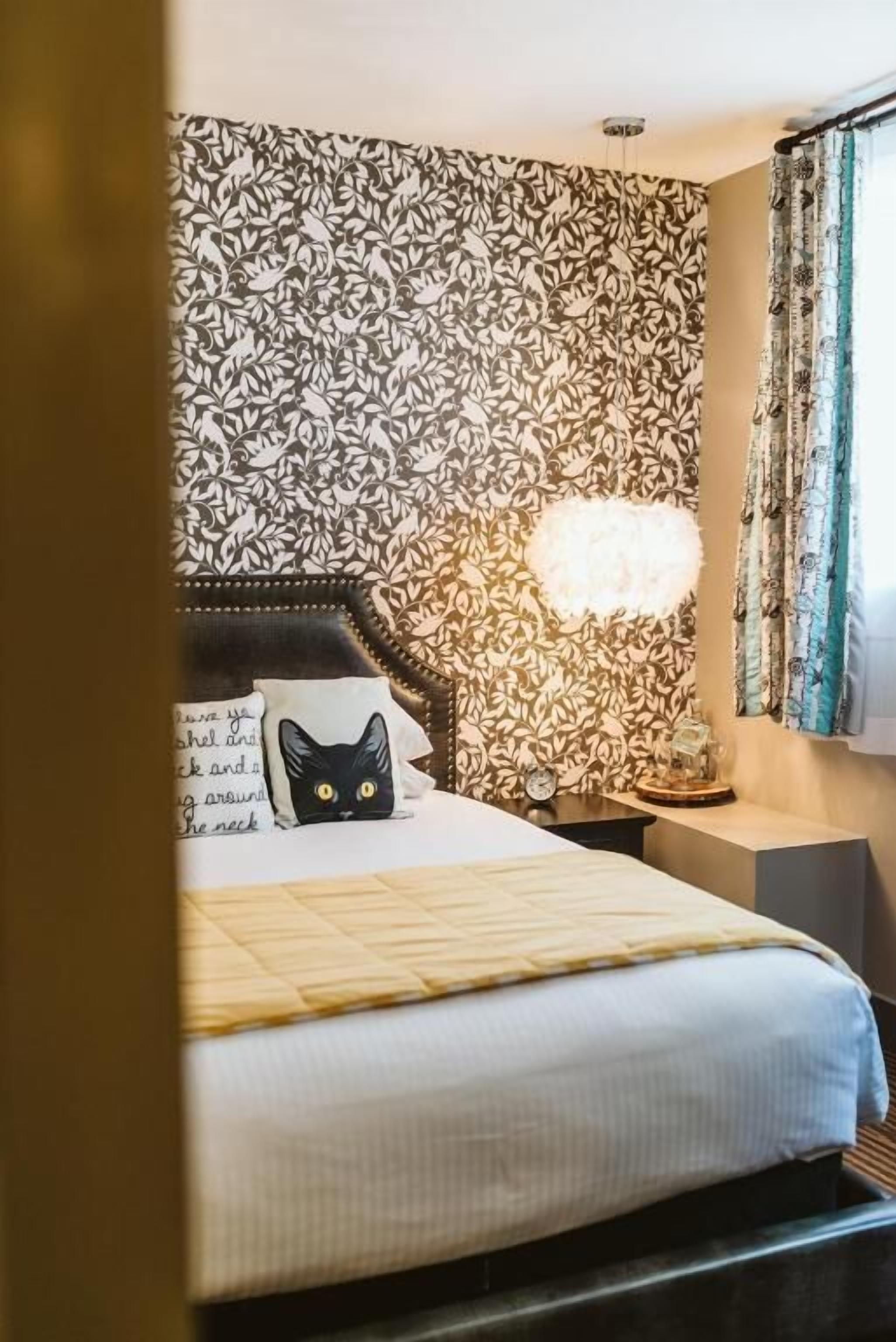 A hotel room with a patterned wallpaper, a bed with a pillow featuring a cat design, a bedside lamp, and a nightstand.