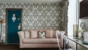 Une pièce avec une porte de couleur turquoise, un papier peint à motifs, un canapé beige, une chaise transparente et une petite table sur laquelle est posé un vase.