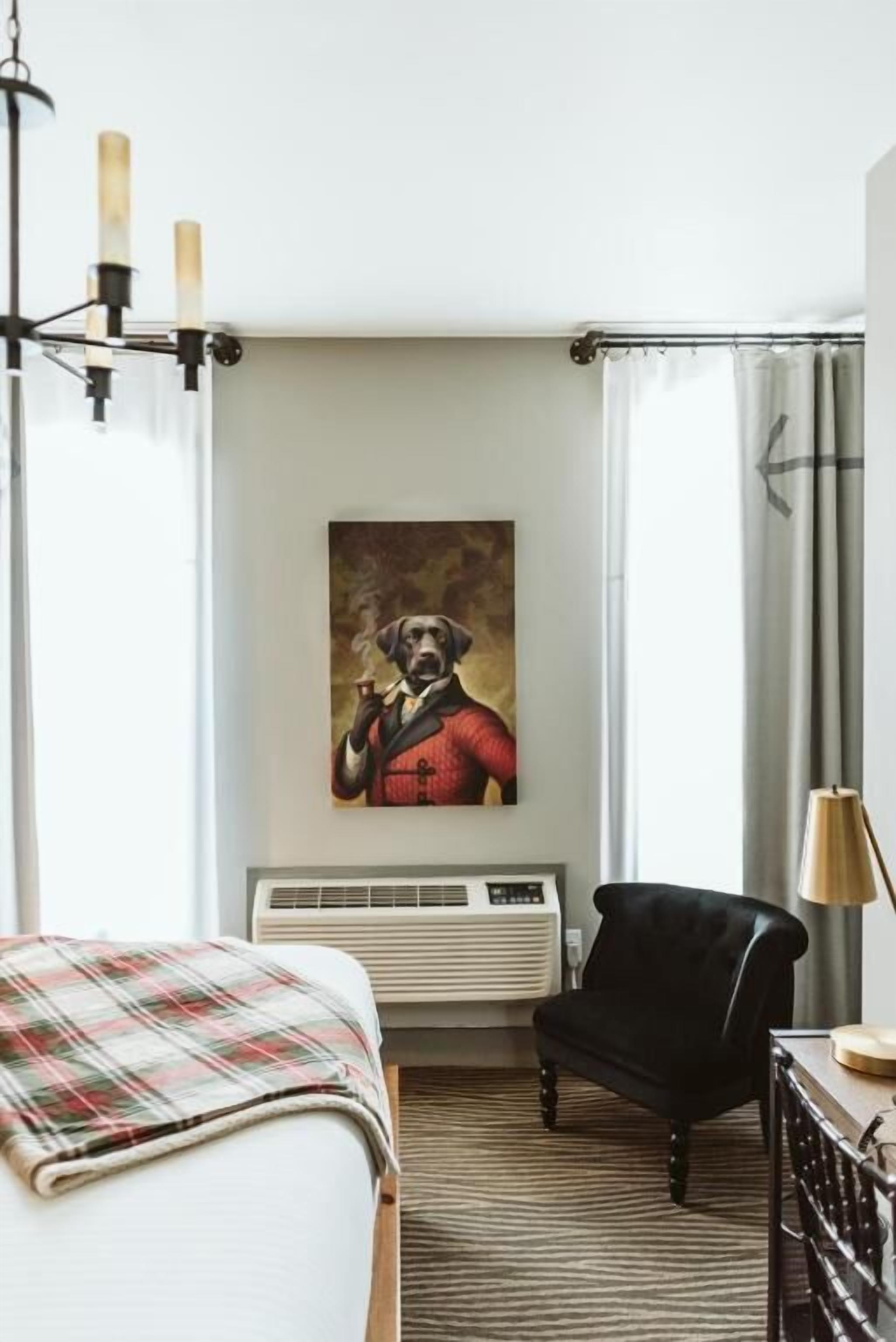 A hotel room with a bed, a chair, a painting of a dog, and a window with curtains.