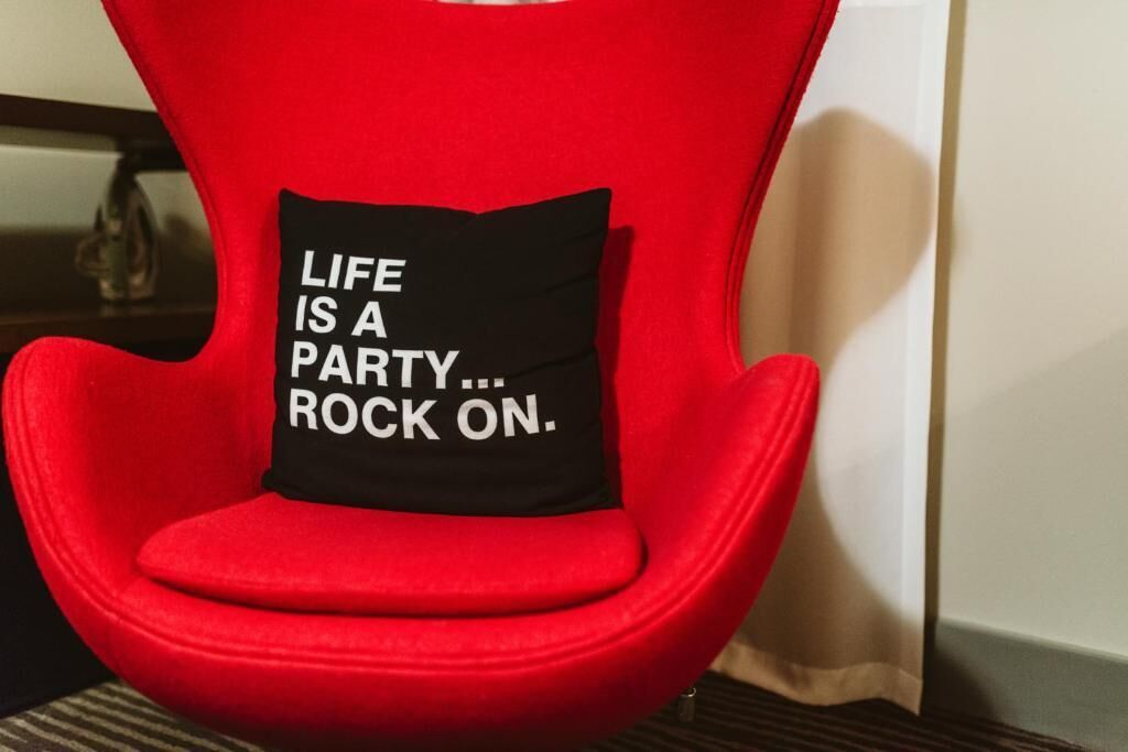 Een rode stoel met een zwart kussen waarop staat 'LIFE IS A PARTY... ROCK ON'.