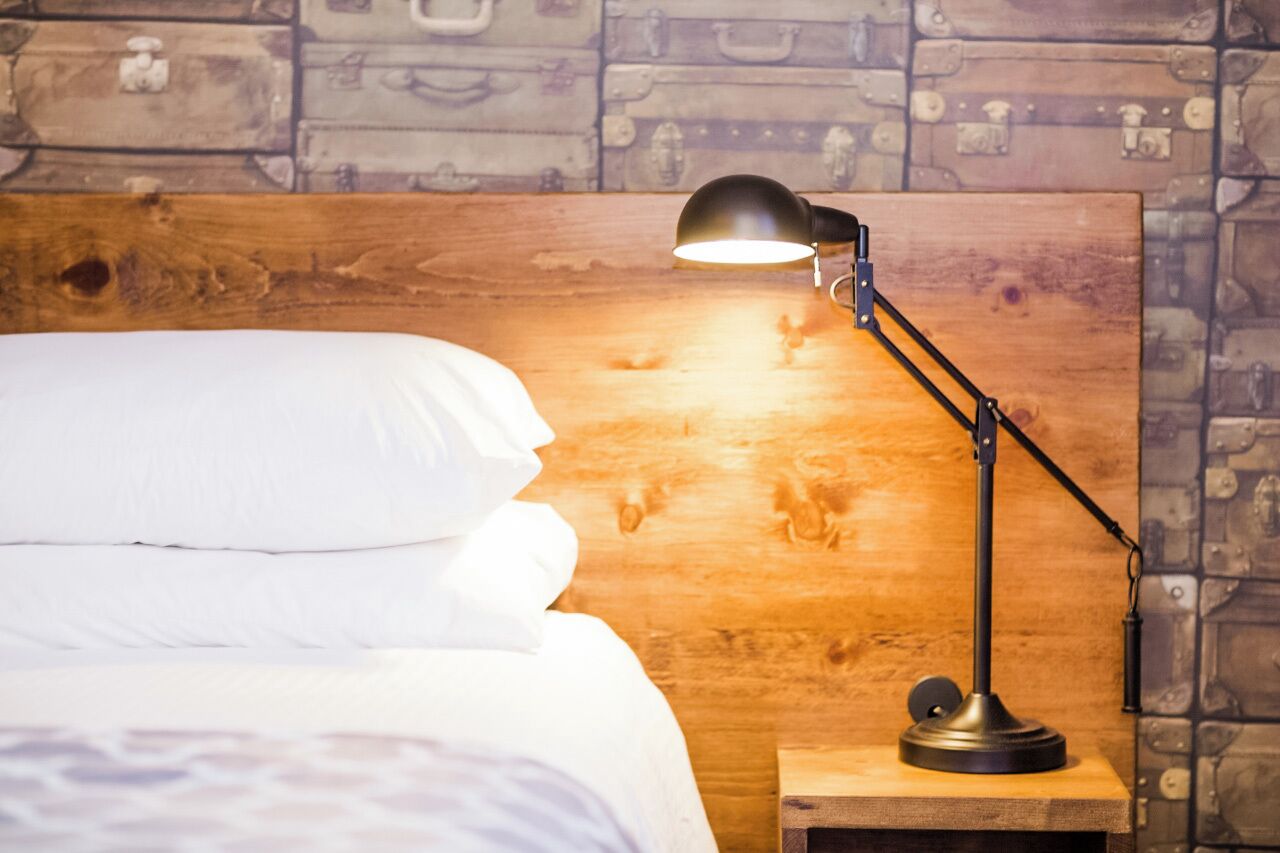 Uma cama com roupa de cama branca, uma cabeceira de madeira e uma luminária de mesa preta sobre um criado-mudo de madeira.