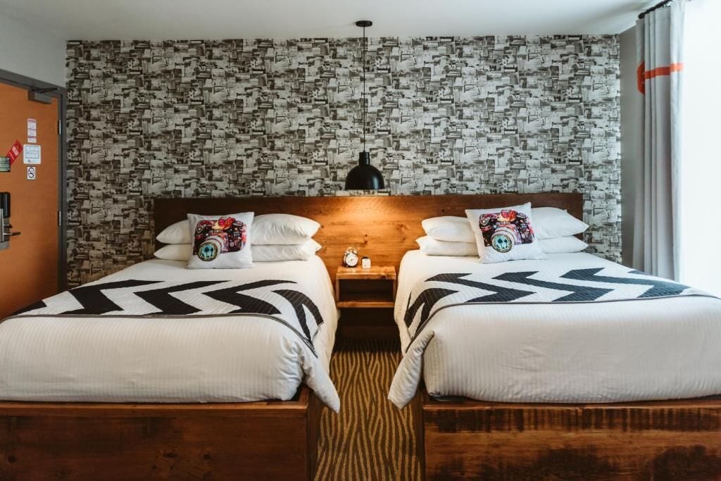 Une chambre d’hôtel avec deux lits, un papier peint à motifs, une tête de lit en bois, une table de chevet avec une horloge et une fenêtre avec des rideaux.