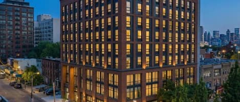 Un immeuble à plusieurs étages, doté d’une façade à fenêtres disposées en réseau, entouré d’autres bâtiments et bordé par une rue.