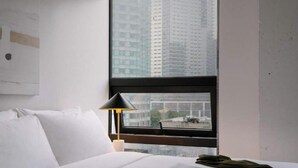 Una camera d'albergo con un letto, un comodino, una lampada e una finestra che si affaccia su edifici alti.