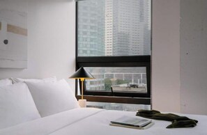 Habitación de hotel con cama, mesita de noche, lámpara y ventana con vista a edificios altos.