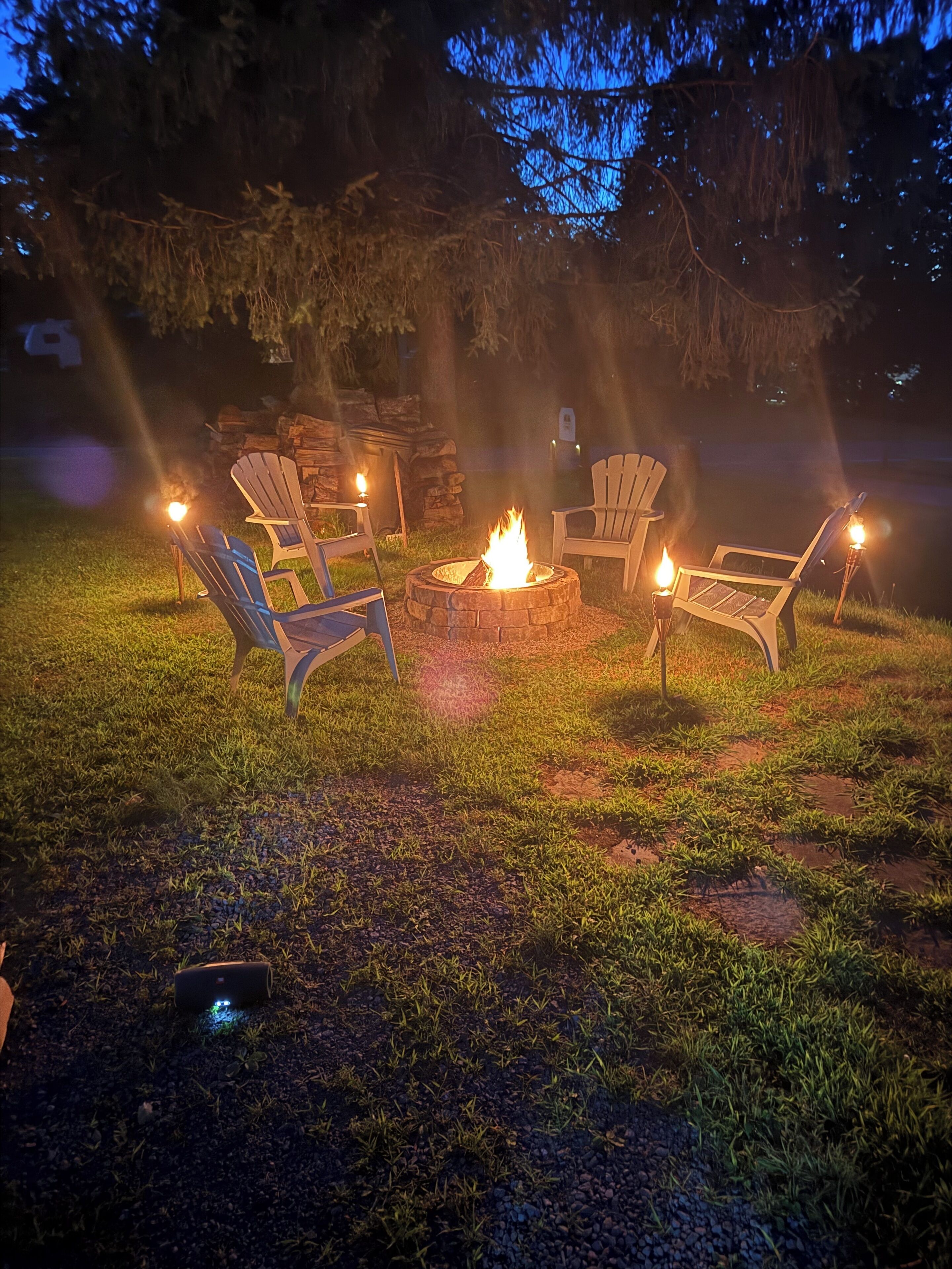 A nighttime outdoor seating area with a fire pit and four chairs, surrounded by trees and a grassy lawn.