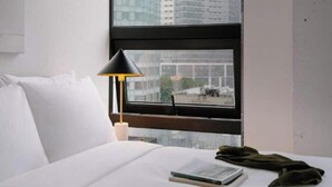 A hotel room with a bed, a window with a view of tall buildings, a black lamp, and a book on the bed.